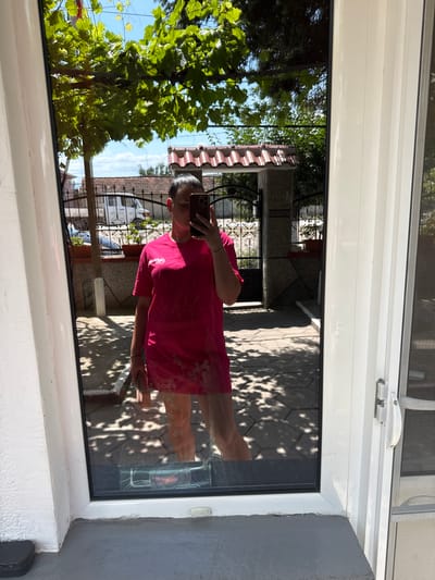 A woman is reflected in a window in Rogosh, Bulgaria, taking a selfie with her phone. She is wearing a bright pink t-shirt dress with a floral pattern at the hem and is holding a pink clutch bag. The background of the reflection shows a courtyard with a paved patio, a fence, and some greenery. The weather appears to be sunny with some dappled shade from trees. The window frame is white, and there is a white handle on the right side of the frame, suggesting it is a door or a large window.