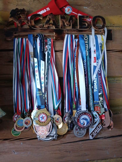 The image shows a wooden display rack mounted on a wooden wall, holding numerous athletic medals. The rack is carved with the word "SAMBO" in large wooden letters. A red banner with "SAMBO-75" is visible at the top, behind the wooden display. The medals hang from the rack by their ribbons, which are a variety of colors including red, white, blue, green, and orange. Many of the medals have inscriptions on them, some of which are partially visible. One medal has "Fight Team Burevestnik" written on it. Another medal has "SAMBO 2022" and "ZVEZDA" visible. The display is situated in what appears to be a rustic setting, possibly a room with wooden walls and floor. The specific location of Verkhnetagirovo, Russia, is not discernible from the image itself. There are no people or discernible actions taking place.