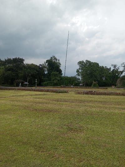 A wide expanse of green grass occupies the foreground, leading to a strip of colorful flowers and low shrubbery in the middle ground. Beyond this, trees and foliage form a dense backdrop. A tall, thin structure, possibly a communication tower or pole, rises above the trees. The sky is overcast with grey, heavy clouds. A small gazebo-like structure is visible to the left, partially obscured by trees.