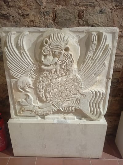 This is a photograph of a relief carving of a winged mythical creature, possibly a griffin, displayed on a stone pedestal in front of a rough-hewn stone wall. The carving is light-colored stone and depicts a creature with a lion-like body, feathered wings, and a head with feline features. The creature's body is detailed with scales or feathers. Its front paw rests on a decorative element. A small informational plaque is visible on the pedestal. The background wall is composed of irregular stones, and the floor appears to be tiled. The carving is illuminated from the front, casting shadows that emphasize its texture and details. The overall scene suggests an exhibit in a museum or historical site.