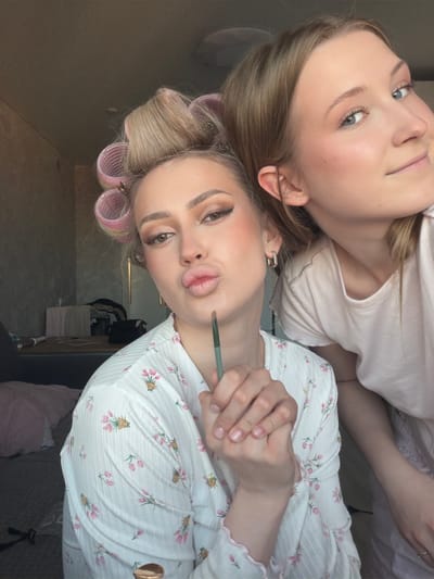 Two young women are depicted indoors in Зубово, Russia, based on the presence of an electronic device on a shelf. The top figure has shoulder-length blonde hair, blue eyes, light skin, and is wearing a light pink top. Her head is tilted, and her expression is neutral. The lower figure has light skin, hair rollers in her blonde hair, and is wearing a white top with a pink floral print. She is pursing her lips in a "duck face" expression and holding a makeup brush near her lips. The background is indistinct, but includes parts of furniture and possibly a towel hanging on a shelf.