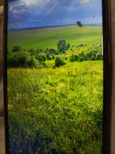 This image depicts a landscape scene on a screen, possibly a smartphone, which exhibits cracks. The scene itself is a vibrant green field under a dramatic sky with clouds. In the foreground, there is tall, lush grass, and further in the background, rolling hills are visible, dotted with trees and what appear to be cultivated fields. The sky is a mix of dark blue and white clouds, suggesting a dynamic weather condition. There are no people or specific urban features visible that would indicate the location as Svoboda, Russia, or any other specific city. The image appears to be a photograph of a natural landscape.