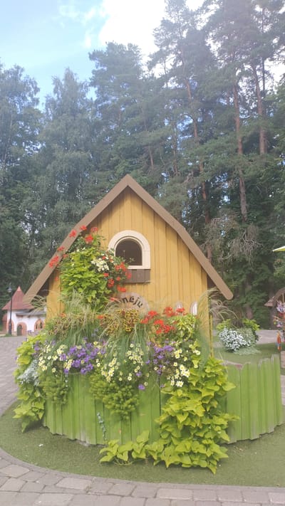 The image depicts a whimsical, yellow, miniature house with a brown roof, nestled amongst lush greenery and flowers. A semicircular window with a cream-colored frame adorns the front of the house. A decorative stone with the engraved word "MĒNEŠU" sits prominently in front of the house, surrounded by a vibrant display of flowers in red, purple, and white, as well as various grasses and leafy plants. A low, bright green, picket-style fence encloses the floral arrangement. To the left, another miniature building with a pointed red roof and white walls is visible. The background consists of a dense forest of tall, mature trees, suggesting a natural setting. The foreground shows a paved pathway made of cobblestones. The scene is illuminated by natural daylight under a partly cloudy sky. This structure is part of the themed park "Lido" located in Kroņauce, Latvia.