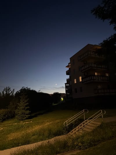 The image depicts an outdoor scene at dusk or night in the city of Lublin, Poland. A multi-story residential building, likely an apartment complex, is visible on the right side of the frame. The building has several balconies with railings and some windows are illuminated from within. In the sky, the moon is clearly visible as a crescent, accompanied by a bright star, possibly Venus. The foreground features a grassy slope with a set of stairs and handrails leading upwards. Trees and bushes are present on the left side of the image, casting shadows. The overall atmosphere is calm and serene, with the natural elements contrasting with the urban architecture.