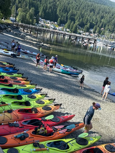 The image depicts a sunny day at a rocky shoreline in the city of North Vancouver, Canada, likely near an area with recreational facilities. A collection of kayaks, in various vibrant colors such as yellow, green, orange, red, and blue, are neatly arranged on the pebble beach, indicating a kayaking or water sports activity. Several individuals, dressed casually in t-shirts and shorts, are present near the kayaks. Some are standing or walking, potentially preparing for or concluding an activity. In the background, a wooden pier extends into the calm water, with sailboats docked alongside it. Beyond the pier, a dense forest covers a hillside, and residential buildings are visible nestled amongst the trees. The scene suggests a popular spot for outdoor recreation and community engagement.