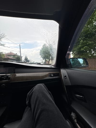 The image, taken from inside a car, presents a rainy day in Градина, Bulgaria. Raindrops cover the driver's side window, partially obscuring the view of houses, power lines, and trees lining the street. The car's interior is visible, including the door panel with a wood trim, a side mirror reflecting the exterior, and a portion of the driver's leg in dark-colored pants. The overall impression is of being inside a vehicle observing a rainy street scene in a residential area of Градина, Bulgaria.