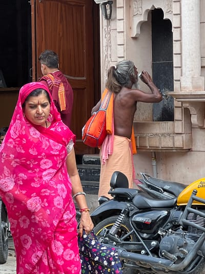 In Pushkar, India, an unclothed man with graying hair tied in a ponytail drinks from a cup while standing near a carved stone structure. He wears an orange cloth draped over his shoulder and a light-colored wrap around his waist. In the foreground, a woman wearing a bright pink floral print sari and matching headscarf walks past. She has a red bindi on her forehead and gold earrings. A motorcycle with a yellow fuel tank is parked nearby. The scene appears to be on a street or in a public area, possibly near a temple or historical site, given the architectural details.