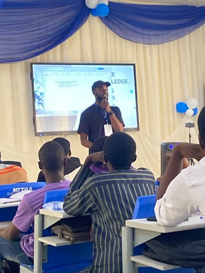 An indoor event is depicted in Baganakwo, Nigeria, featuring a male speaker addressing an audience. The speaker, positioned centrally, is a dark-skinned male with a beard, wearing a black t-shirt, black baseball cap, and a white lanyard. He holds a black microphone to his mouth with his right hand. Behind him is a large flat-screen display projecting a presentation slide with the word "KNOWLEDGE" prominent on the right side. The left side of the screen shows what appears to be a document or slide content, partially visible as "Lorem ipsum dolor sit amet" and "Wellington boot 2023." The room's upper portion is decorated with blue and white fabric drapes and clusters of blue and white balloons.
The audience, visible primarily from behind in the foreground, consists of multiple individuals seated at blue and white school-style desks. One individual wears a purple t-shirt, another a vertical blue and yellow striped long-sleeve shirt, and a third a white shirt. Water bottles and various papers are visible on the desks. A speaker unit is present on the right side of the stage area.
