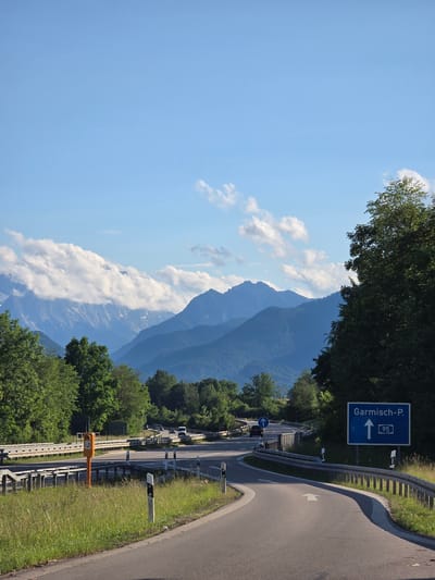 This image depicts a highway interchange near Ohlstadt, Germany, under a clear to partly cloudy blue sky. A panoramic view of the Bavarian Alps dominates the background, with multiple mountain ranges receding into the distance, exhibiting atmospheric perspective. The nearest mountains are deep blue-green, covered in dense forest, while the more distant peaks on the left show snow patches and are partially obscured by a thick band of white cumulus clouds.

In the mid-ground and foreground, a complex road junction is visible. The main road curves sharply to the right and ascends, with a smaller road branching off to the left and curving downward. A prominent blue rectangular road sign on the right indicates "Garmisch-P." (Garmisch-Partenkirchen) with an upward arrow and the route number "95" (Bundesautobahn 95).

Multiple vehicles, including several dark cars and a white van, are visible on the roads in the mid-ground, heading away from the viewer. Metal crash barriers (guardrails) line the edges of the roads, which are marked with white lane lines and reflective white posts with black tops. In the central-left foreground, an orange pole with a sensor-like component stands between the diverging road sections. Two empty black rectangular signs are positioned further back on the left, above the orange pole. A smaller blue circular sign with a white arrow indicating mandatory straight direction is also visible on the left road section.

The road surfaces are asphalt, and the verges are covered in green grass and wildflowers, particularly on the left side, where tall grasses are present. Deciduous trees with vibrant green foliage line both sides of the roads and cover the lower slopes of the mountains, indicating a lush, natural environment. The scene is illuminated by bright sunlight, casting distinct shadows, suggesting mid-morning or late afternoon. No individuals are visibly present.
