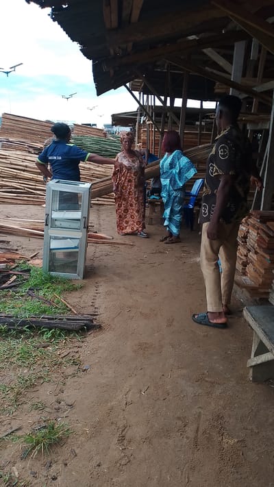 A medium-shot image captures a scene in Asubi, Nigeria, possibly a lumber or construction site. The ground is earthen, with scattered vegetation and debris. A stack of bamboo and lumber is visible in the background, partially covered by a makeshift wooden structure. Five people are present: one in a dark blue shirt marked "SANITATION TEAM," gesturing with their right arm; a woman in a brown and orange patterned dress and headwrap; a woman in a blue patterned dress; a man in khaki pants, a dark patterned shirt, and sandals, standing with his back mostly to the camera; and another woman that is partly behind the first woman. To the left of the "SANITATION TEAM" member is a small, free-standing aluminum and glass cabinet. Beside the man is a stack of wood.