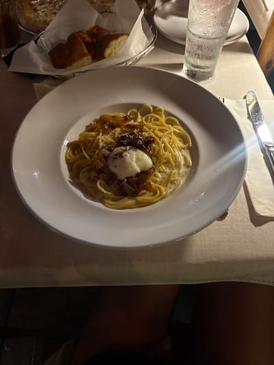 The image shows a plate of pasta with a creamy sauce, topped with a white ball (possibly cheese or burrata) and some darker pieces (likely meat or vegetables), set on a white tablecloth. Beside the plate, there's a glass of water with condensation and a basket of bread. The utensils are placed on a napkin above the plate. The scene appears to be at a restaurant, given the presentation of the food and the table setting. Though the exact restaurant or location in Honolulu is not discernible from the image, the style of food suggests it could be an Italian or European-influenced establishment.