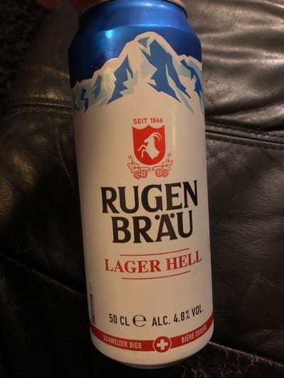 The image shows a horizontally oriented can of Rugen Brau Lager Hell beer against a dark, possibly leather, textured surface. The can is white with blue detailing around the top edge. The can displays the brand "Rugen Brau" in black, with the words "Lager Hell" in red beneath it. The can's labeling also indicates "50 CL e" and "ALC. 4.8% VOL." and contains the Swiss flag with the text "SCHWEIZER BIER BIERE SUISSE". It also bears the mark "SEIT 1866". There are no people visible in the image. Rugen Brau is a brewery located in Interlaken, Switzerland, not Kehrsatz.