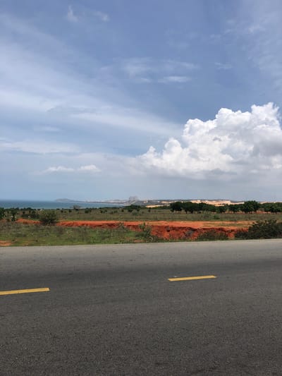 This is a vertical, eye-level shot of a coastal landscape in Hòa Thắng, Vietnam, captured from the side of a paved road. The foreground is dominated by the asphalt road, marked with yellow lines. Beyond the road, a strip of orange soil and green vegetation stretches horizontally. Further back, a line of dark green trees stands against a backdrop of sand dunes and distant buildings. The sea is visible to the left, meeting a hazy horizon under a sky filled with white and gray clouds. There are no people or animals in the frame.