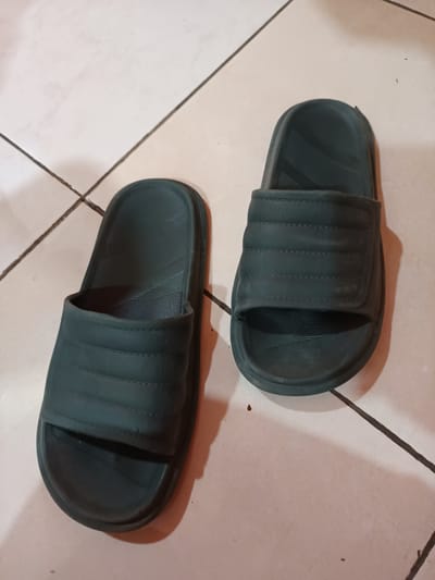 This is a top-down, eye-level shot of two dark green slide sandals placed on a tiled floor. The sandals are positioned diagonally on the floor, with the left sandal slightly closer to the viewer. Both sandals are identical, featuring a broad strap across the top with three horizontal stitched lines. The soles are textured with parallel lines. The floor is made of light-colored square tiles with dark grout lines. The lighting appears to be artificial, casting soft shadows. There are no people visible, and the sandals are the sole focus of the image. The setting appears to be indoors, possibly a home or a changing room. The overall impression is casual and domestic.