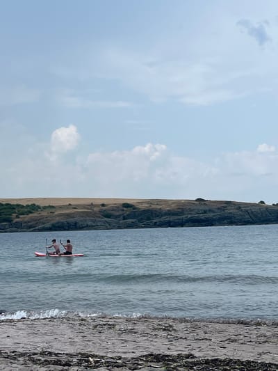 In Ahtopol, Bulgaria, two individuals are paddleboarding on the sea. They are positioned on a single paddleboard, with one person in front and the other behind. Both are actively paddling. The water is calm with small waves. In the background, a rocky coastline with dry, grassy hills rises from the sea. The sky is partly cloudy. In the foreground, there is a beach with sand and scattered debris.