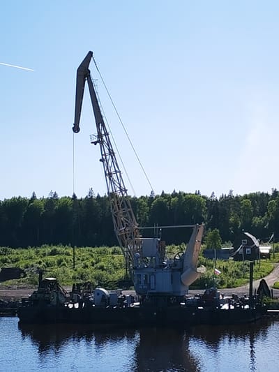 This image depicts an industrial scene featuring a large, older model crane mounted on a dark-hulled barge or floating platform in a body of water. The crane has a prominent lattice boom extending towards the upper left, with a solid, angled jib. Cables and rigging support the boom. To the left of the main crane, another piece of heavy machinery is visible on the platform, suggesting an industrial or dredging operation.
In the background, a dense line of green trees forms a forest, stretching across the horizon. On the right bank, a small, dark-roofed building with white windows is partially visible amidst the foliage. A small flag, clearly identifiable as the Russian flag with its white, blue, and red horizontal stripes, is planted near this building. The foreground shows the calm, reflective surface of the water, mirroring the platform and parts of the sky.
The scene is illuminated by bright daylight under a clear, pale blue sky, indicating sunny weather. A thin, white contrail from an aircraft is visible in the upper left corner of the sky. There is no visible activity or people. The overall impression is one of a quiet industrial site nestled within a natural, wooded landscape. Faint, illegible text might be present on the crane's cabin. The location context places this scene in Davydovskaya, Russia.