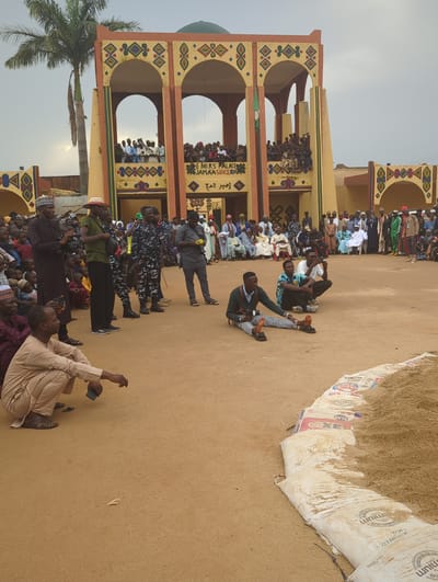 A partially-rotated outdoor scene in Kafanchan A, Nigeria, shows a crowd of people gathered near a three-story building with archways, with more people visible on the building's levels. The building is painted in yellow and brown with decorative patterns. Some people are standing, while others are seated on the ground. Law enforcement personnel in camouflage uniforms are present in the foreground. There are large bags of grain or other material to the right of the scene. A palm tree stands to the left.