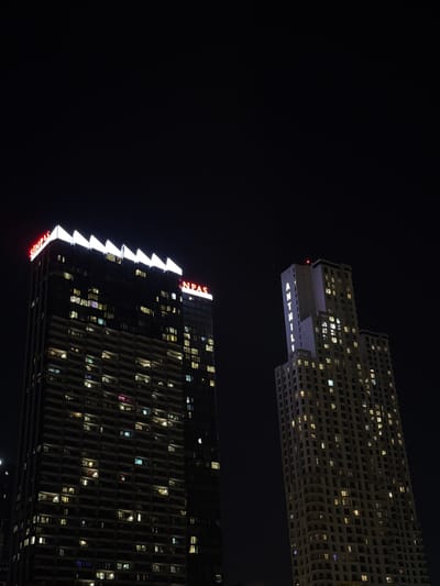Two modern buildings are silhouetted against a dark night sky. The building in the foreground is a tall, dark skyscraper with many windows illuminated from within, showing hints of various colored lights. The building has a distinctive jagged, white illuminated edge running down its side, and red neon signage spelling out "SINPAS" at the bottom and top. The building in the background, which appears to be farther away and possibly higher, is lighter in color and also features many illuminated windows. It has a distinctive, stepped architectural design and text that appears to read "ANTELIA" in white illuminated letters on its lower facade. The scene suggests an urban setting at night, possibly in Şişli, Turkey, given the location context. There are no people visible, but the lit windows imply occupancy and activity within the buildings. The overall mood is one of nighttime urban vibrancy and architectural presence.