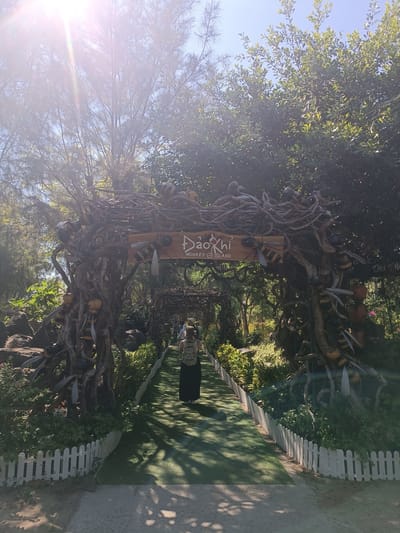This media file depicts an outdoor scene with a pathway leading through a decorative archway adorned with large, stylized bee figures and intertwined branches. The archway is made of natural materials, appearing to be crafted from twisted roots and branches. The path itself is a vibrant green, possibly artificial turf, bordered by a low white picket fence.
A person, seen from behind, is walking along the path away from the viewer, wearing a backpack and a long dark skirt or dress. Other individuals are visible further down the path, also walking. The setting is lush and green, with abundant trees and foliage surrounding the pathway and archway, suggesting a park or natural attraction. The time of day appears to be morning or afternoon, with bright sunlight streaming through the trees, casting dappled shadows on the path. The sky is clear and blue.
A sign is visible on the archway with text that reads "ĐẢO KHỈ MONKEY ISLAND." This indicates the location as Monkey Island. The overall atmosphere seems serene and inviting, drawing visitors into a themed environment. The large bee decorations add a whimsical and playful element to the natural surroundings. The light flare in the upper left corner suggests a sunny day.