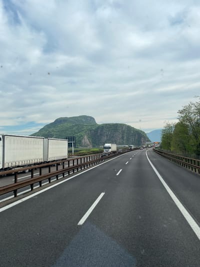 The image shows a view from a vehicle traveling on a highway. The road has two lanes marked by white lines. A rusty metal guardrail separates the highway lanes from a parallel service road or shoulder where several semi-trucks are traveling. Beyond the service road is a mountain covered in green vegetation. The sky is overcast with a mix of white and gray clouds. The location is near Tramin an der Weinstraße - Termeno sulla Strada del Vino, Italy, based on the mountainous landscape and general location. There is a line of vehicles in the distance on the highway.