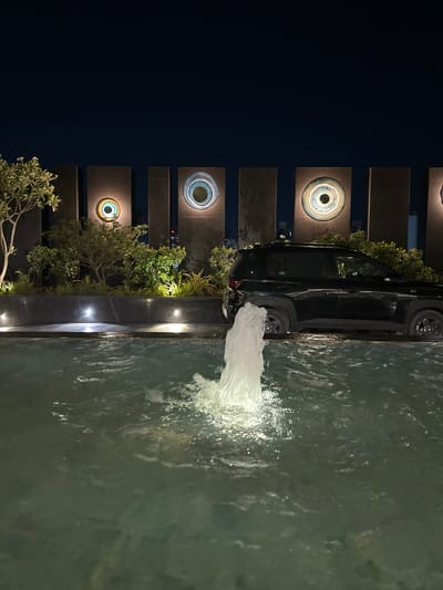 The image shows an outdoor water feature at night, likely in Vadodara, India. A dark-colored van is parked alongside the feature. The feature consists of a wall with decorative circular elements interspersed with greenery. Water is being pumped into the feature, creating a visible splash. The lighting is artificial, with some lights illuminating the water and the wall. The scene appears to be a modern architectural design, possibly part of a hotel or a high-end residential complex.