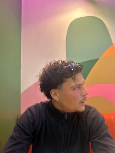 A person with dark, curly hair is looking up and to the right. They are wearing a dark gray zippered jacket and an earring in their left ear. The background consists of a large, abstract mural with overlapping pastel colors, including pink, green, orange, and yellow. The colors are in irregular, curved shapes. The location is likely indoors, in Serris, France, given the request.