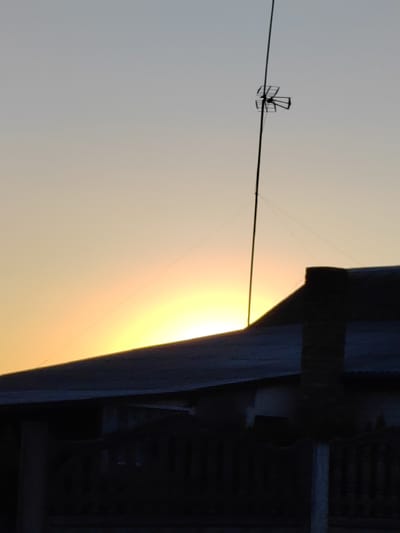 This image depicts a silhouetted rooftop and fence against a pale orange and yellow sky, suggesting either sunrise or sunset. A tall, thin antenna with a directional element at its top stands prominently against the sky. The sky itself is a gradient, transitioning from a lighter hue at the top to brighter, warmer tones near the horizon. The rooftop is dark and appears to be corrugated metal. A brick chimney or structure rises from the roof. In the foreground, a dark, silhouetted fence with vertical posts is visible. The overall scene evokes a quiet, possibly rural setting. The light suggests the early morning or late evening. The provided location context of Воўчынскі сельскі Савет, Belarus, places the scene in a specific geographic region. There is no visible text in the image.