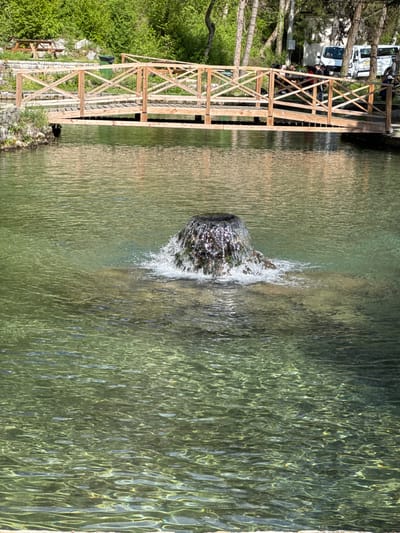 A landscape photograph shows a wooden bridge next to clear water in Milanovo, Bulgaria. The bridge stands to the left of the frame. A small fountain or water feature is located in the center of the water, creating a splash effect. The water is so clear that the bottom can be seen. Trees and vegetation are visible on the left side of the bridge. Some vehicles are also visible in the background on the left side. The scene is bathed in sunlight.