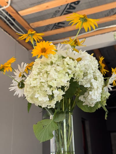 The image shows a close-up of a bouquet of flowers in a glass vase. The bouquet is composed of white hydrangeas, yellow daisies, and some other white flowers with yellow centers. Some green leaves are also visible. The flowers are arranged in a way that they appear to be cascading downwards. The vase is clear and contains water and stems.
In the background, there is a construction site, suggested by wooden beams and what appears to be building insulation material. The insulation has some text printed on it, but it is partially obscured and not entirely legible. The scene is brightly lit, suggesting daytime. The overall impression is one of a natural arrangement set against an unfinished, industrial backdrop. There are no people visible in the image. The location context is Novotitarovskaya, Russia.