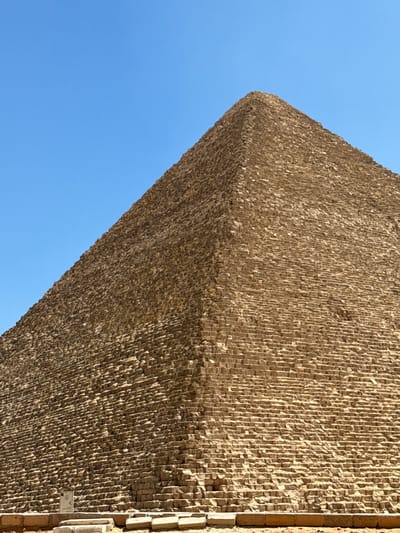 The image shows a close-up, low-angle view of the Great Pyramid of Giza, also known as the Pyramid of Khufu, in Giza, Egypt. The pyramid's rough, massive stone blocks dominate the frame, with their weathered textures clearly visible. The pyramid's steep slope rises towards a sharp apex, meeting a clear, bright blue sky. The bottom of the image reveals the base of the pyramid and a few flat stone slabs and some rubble on the ground in front of it. A small, white informational sign with some text and symbols is visible near the bottom left of the pyramid's base. The scene is devoid of any people or animals.
