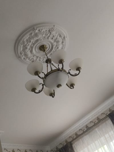 An indoor shot captures a traditional chandelier hanging from a white ceiling adorned with ornate plasterwork. The chandelier features multiple frosted glass shades and a brass-toned metal frame. Below the ceiling, a decorative cornice with a repeating floral pattern is visible, running along the top of a wall. A window with sheer curtains and darker drapes is also present in the lower right of the frame. The scene appears to be in a residential or formal setting in Мещерино, Russia. There are no people or animals visible in the image.