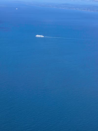 An aerial shot captures a vast expanse of deep blue ocean under a hazy sky. A large white cruise ship is visible in the mid-right section of the image, leaving a white wake as it sails. Further in the distance, to the left of the cruise ship and closer to the horizon, a smaller white vessel is also present. The distant horizon shows a faint outline of land, possibly the coast of Ireland, with some buildings visible along the shore. The water exhibits subtle ripples and undulations, indicating a calm sea. The image does not contain any specific details that can be definitively linked to Dublin, Ireland, other than the potential distant landmass.