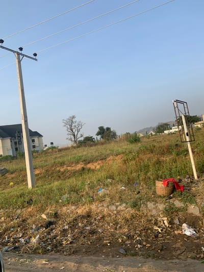 A grassy field meets a dirt path, marred by scattered trash. A wicker basket with a red cloth sits in the field. Buildings appear in the distance, along with trees. A utility pole stands tall, wires stretching into the clear sky. The scene suggests a blend of rural and developed environments.