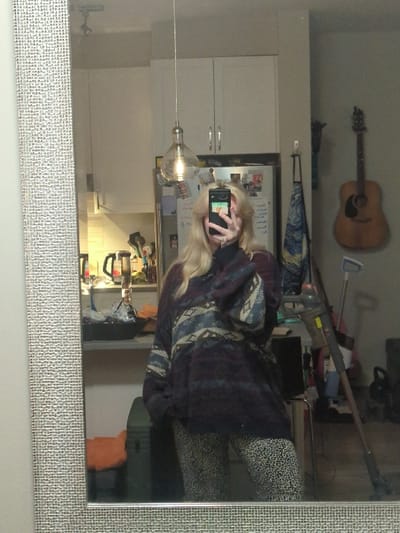 The image depicts an indoor scene, observed through a mirror reflection, stated to be located in Area D (Elaho/Garibaldi), Canada. A single individual with long blonde hair, whose face is obscured by a dark-colored smartphone held in their right hand, is positioned centrally, capturing a self-portrait. The individual wears a loose-fitting, patterned pullover sweater featuring geometric designs in shades of dark purple, blue, beige, and maroon, paired with animal-print leggings or pants. A ring is visible on the fourth finger of their left hand.
The background, viewed in reflection, presents a domestic environment. To the viewer's left, a kitchen area is visible, containing white upper cabinets with silver handles, a stainless steel refrigerator adorned with multiple magnets, and a counter holding items including a black bowl, a kettle, and various utensils. A clear glass pendant light with an exposed filament bulb hangs from the ceiling in the mid-ground. An orange fabric item is partially visible on a lower surface in this area. To the viewer's right, an acoustic guitar with a light wood finish hangs on a neutral-colored wall, alongside a blue, white, and yellow patterned fabric suspended from a hook. A cordless stick vacuum cleaner leans against a wall or furniture to the right of the individual, and a dark green storage container is visible on the floor. The mirror itself features a textured, silver-colored frame.