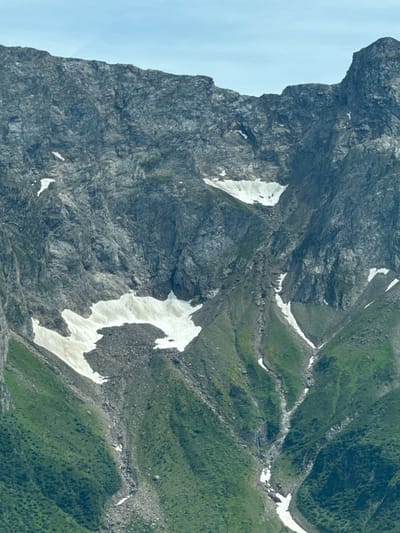 This media file is a landscape photograph taken from an aerial perspective of a steep, rocky mountainside with patches of snow and verdant green vegetation. The scene is dominated by the rugged texture of the rock face, characterized by its grey and dark tones, with deep crevices and uneven surfaces. Several distinct patches of white snow are scattered across the rocks, indicating a high-altitude environment where snow persists even in warmer seasons.
Interspersed with the rock and snow are slopes covered in lush green grass and low-lying vegetation. These grassy areas are not uniformly smooth but appear to be textured with small shrubs or tufts of grass, creating a dappled effect. A narrow, winding path or a dry streambed can be faintly discerned traversing the landscape, connecting different sections of the mountainside and bordered by both rock and vegetation.
The lighting suggests it is daytime, with the sun illuminating parts of the landscape, creating shadows that accentuate the topography. The sky is a pale, overcast blue, visible on the left side of the frame, hinting at potentially cool weather or simply a diffused light source. There are no people or man-made objects visible in the frame, emphasizing the wild and natural state of the environment.
The overall impression is one of a remote, mountainous terrain, likely part of the Swiss Alps given the location context of Airolo, Switzerland. The combination of rock, snow, and green slopes creates a visually striking contrast and highlights the dynamic nature of alpine landscapes. The image captures the raw beauty and scale of the mountains, evoking a sense of ruggedness and natural grandeur.