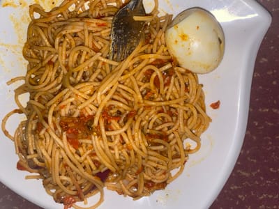 A white, irregularly shaped plate, positioned on a reddish-brown patterned surface, contains a portion of spaghetti coated in a reddish-orange sauce with visible chunks of tomato and possibly other ingredients. A silver-colored metal fork is partially embedded in the spaghetti, tines down, near the upper center. To the upper right, a peeled, hard-boiled egg with minor sauce residue is resting on the plate. No individuals are present in the scene, which is located in Mape, Nigeria.