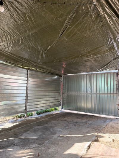 The image depicts the interior of an unfinished or partially constructed structure, likely a garage or covered workspace, located in Pňovice, Czechia. The space is defined by corrugated metal walls and a large tarp serving as a temporary roof. The ground is rough concrete, showing signs of recent work or unevenness, with some sand and debris present. Sunlight casts long shadows across the floor, indicating an outdoor or open-air setting. The walls appear to be made of vertical corrugated metal panels, with one section a gate-like structure, possibly a garage door, partially open. A string of small lights is visible along the underside of the tarp. The overall impression is one of a functional, work-in-progress environment. There are no people or animals visible in the image.