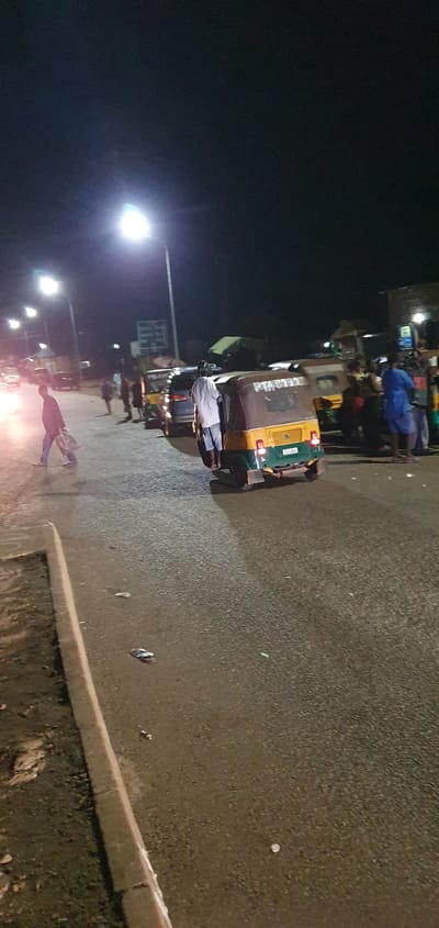 At night in Barakin Angundi, Nigeria, a street scene unfolds under the glow of streetlights. The asphalt road is wet, reflecting the lights and creating a shimmering effect. Several vehicles are present, including a yellow and green tuk-tuk in the foreground with visible writing on its side. A dark-colored van is parked behind it. In the background, more vehicles and various structures line the street, suggesting a commercial or transit area.

People are scattered across the scene. A man wearing a white shirt and blue shorts stands near the tuk-tuk. Further to the left, a person in a light-colored outfit walks across the road, carrying bags. Other individuals are visible in the background, some standing near the vehicles and structures, their actions unclear in the low light. The overall atmosphere is that of a busy street at night, possibly a transportation hub or market area, illuminated by artificial lighting. The presence of debris on the wet road indicates recent activity or potential neglect.