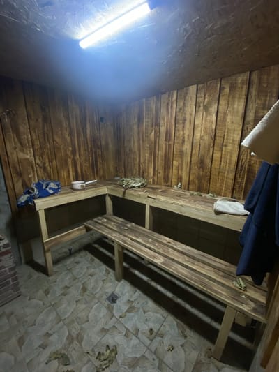 The image depicts the interior of a wooden sauna. The walls and benches are constructed from rustic, dark-stained wood. A fluorescent light fixture illuminates the space from the ceiling, which appears to be paneled with oriented strand board (OSB). On the benches, there are several items: a pile of dried branches, possibly for use as a venik (a sauna whisk), a small metal pot with a handle, a blue and white patterned cloth, and a light-colored towel. A dark blue garment is hanging on the right side of the image. The floor is tiled with large, irregularly shaped tiles in shades of beige and grey. Scattered on the floor and benches are dry leaves. In the lower left corner, a section of brick wall is visible. A drain grate is present in the tiled floor. The scene is devoid of any people. This sauna appears to be located in Kisliakovskaya, Russia, given the context provided.
