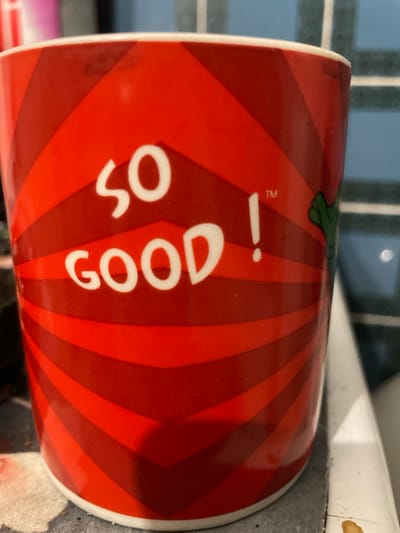 A close-up photograph captures a cylindrical mug, positioned on a speckled gray countertop in what appears to be a kitchen or utility area within Senden, Germany. The mug is predominantly red with a repeating pattern of alternating bright red and darker red diagonal stripes, forming a chevron-like design around its circumference. Prominently displayed on the front of the mug in white, stylized capital letters is the phrase "SO GOOD!", with a small superscript "™" symbol located above and to the right of the exclamation point. A partial green, cartoon-like appendage, possibly a hand or tentacle, is visible emerging from the right side of the mug's design. The mug has a white rim at its top and bottom. The background features light blue rectangular wall tiles with white grout lines. A white vertical surface, possibly the edge of a sink or appliance, is visible to the right of the mug. A dark, irregularly shaped stain is present on the countertop surface to the lower right, near the base of the white vertical surface. No people are visible, and no explicit actions or interactions are depicted.
