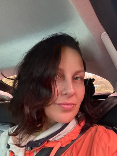 A close-up, selfie-style, medium shot captures a woman in the backseat of a vehicle. She has dark, wavy hair that falls around her face and shoulders. Her gaze is directed slightly away from the camera, with her left eye, a green hue, being more visible. Her lips are slightly parted and appear to be a pale pink. She is wearing an orange jacket over a white and dark-striped garment. A black seatbelt is visible across her chest and shoulder. The background shows the interior of a car, with a gray headliner and a window revealing a blurred outdoor scene with trees. The specific location is within the city of Gremyachinsk, Russia.