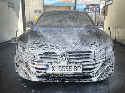 The image depicts a gray Volkswagen Arteon sedan undergoing a wash at a car wash facility. The entire front of the car, including the hood, windshield, front grille, and bumper, is covered in white soap suds. The car has a Bulgarian license plate that reads "K 5555 BP". The car wash bay has concrete walls. On the left wall, there is a sign written in Cyrillic that reads "ТУРБОПЯНА". On the right wall, there is a sign that reads "ПРАХОСМУКАЧКА ЕДНОПОСТОВА" and advertising posters for autobox.bg, Duracell Automotive batteries, and other automotive products. Based on the license plate prefix "K", the location is likely in Krumovgrad, Bulgaria.