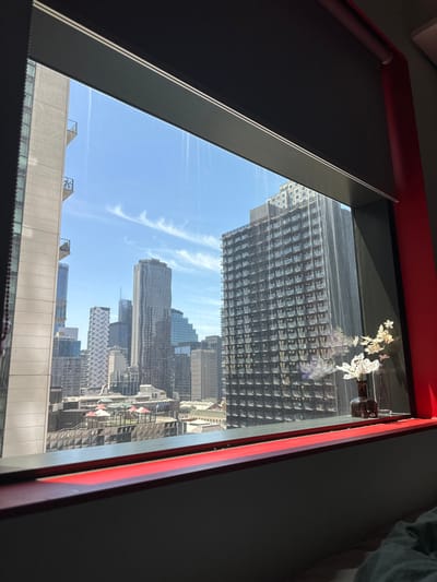 A partial view from a window reveals a cityscape. The sky is blue with wispy clouds. Skyscrapers dominate the horizon, exhibiting diverse architectural styles and heights. Some are tall and gridded, others more sleek and modern. The lower portion of the view includes closer buildings with visible rooftops and structures. A red window frame surrounds the scene. A dark window blind is partially drawn and some white flowers are set on the windowsill.