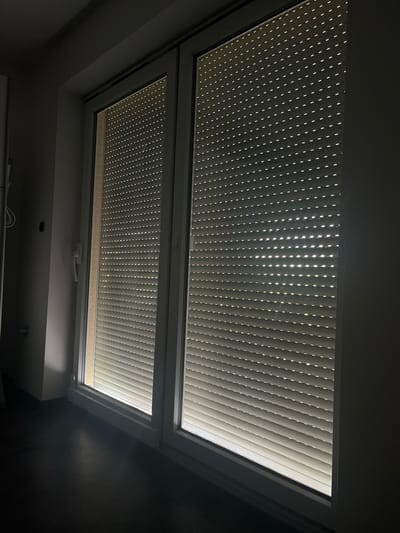 The image shows a dark interior scene of a room with a large window covered by rolling shutters. The shutters are partially open, allowing light to filter through small horizontal slits, creating a striped pattern of illumination across the dark surfaces. The window appears to be a double-pane sliding door with a white frame. To the left of the window, a white wall is visible, with a light fixture and a portion of a cable. The floor is dark and reflective, suggesting it is made of polished wood or a similar material. The overall impression is one of a dimly lit, modern living space. The scene does not provide any specific indicators of its location within the city of Siegen, Germany, as it lacks any recognizable landmarks or identifying features.