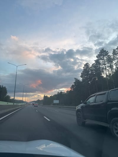 The image shows a view from the perspective of a vehicle driving on a highway. The highway has multiple lanes and is bordered by trees on the right and a concrete barrier on the left. Several cars are visible on the highway, including a black pickup truck in the foreground to the right. The sky is cloudy with hints of pink and orange, suggesting either sunrise or sunset. Streetlights are present along the highway. The environment appears to be a natural landscape adjacent to a major roadway. The location is not specified as Kapan', Russia.