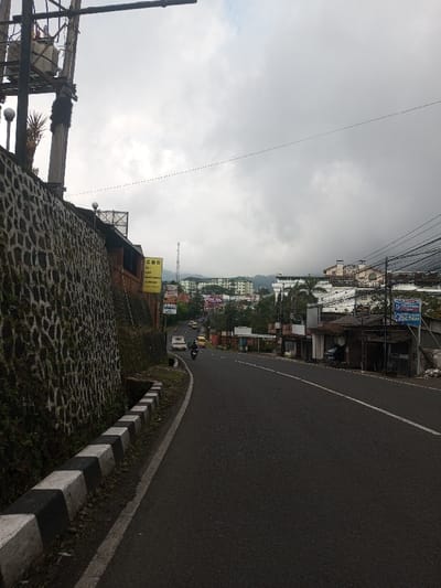 A road curves through a hillside town under a cloudy sky. A tall stone wall runs alongside the road, marked with a black and white curb. Buildings line the opposite side of the road, interspersed with power lines and signs. In the distance, taller structures can be seen nestled in the hills. Traffic moves along the road, including cars and motorbikes. A utility pole stands prominently on the left side of the frame.