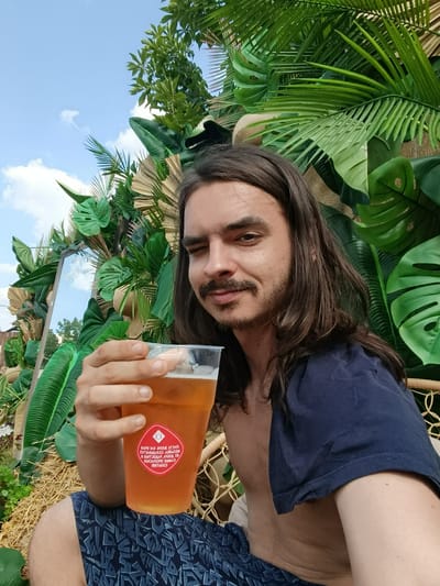 A young man with long brown hair and a mustache takes a selfie while holding a plastic cup of beer. He is winking at the camera and appears to be enjoying himself. He is wearing a dark blue t-shirt and patterned shorts. The background is a lush display of tropical plants, including large palm leaves and other greenery, with a mirror reflecting the sky and some trees. The lighting suggests it is daytime, with a bright, clear sky and some scattered clouds. The overall scene evokes a festive or relaxed outdoor atmosphere. On the cup, there is a red circular sticker with white text, which appears to be in Cyrillic. The text on the sticker reads "ВЯЧЕСЛАВ АЛЕКСАНДРОВИЧ" and "КОРМЛЕНИИ" among other illegible text.