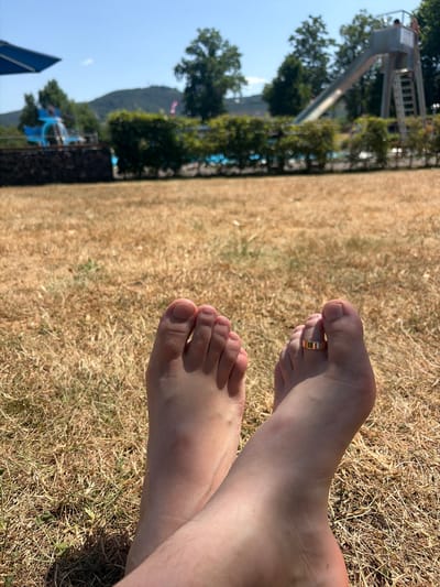 The image shows a close-up view of two feet resting on dry grass. One foot is positioned slightly in front of the other, and a gold ring is visible on the second toe of the foot in front. The feet appear to be bare and have fair skin. The background is a blur of dried grass, suggesting an outdoor setting. In the distance, there are hints of trees and possibly a hillside, with a bright blue sky visible. The lighting indicates it is daytime, likely sunny.
Further elements visible in the background, though out of focus, include what appears to be a playground structure with a slide and ladder, and possibly some water features. The presence of these elements hints at a recreational area, such as a park or an amusement area. The overall impression is one of relaxation and leisure. No text is visible in the image.