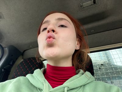 The image depicts a young adult female seated inside a vehicle, captured in a close-up, selfie-style perspective. The subject has reddish-brown hair and fair skin. Her lips are puckered into a "kiss" pose, directed towards the camera. She wears a red turtleneck shirt layered under a light green hooded sweatshirt. A small, round, silver-colored stud earring is visible in her left earlobe.
The interior of the vehicle shows a light gray fabric headliner with an overhead light fixture and a circular cap or vent. A black seat or seat cover with a red diamond-stitched pattern is partially visible behind the subject. To the right of the subject, a window with a light-colored, metallic grid or mesh pattern covers the glass, through which an indistinct, overcast exterior is visible. The precise location within Siktarva, Georgia, cannot be determined from the image.