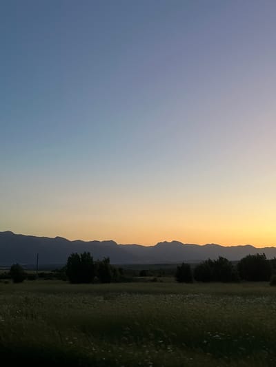 The image shows a landscape at sunset in Gorna Kremena, Bulgaria. The sky transitions from blue on the left to orange/yellow towards the right. A mountain range is silhouetted against the sunset sky. The foreground is a field with scattered plants, and some trees are visible. A single utility pole is also visible near the bottom of the image. The image is rotated 90 degrees counter-clockwise.
